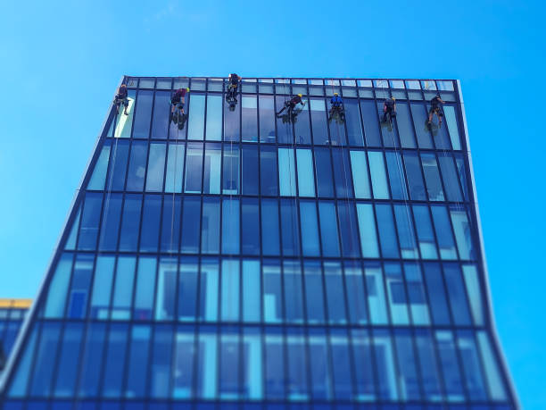 Rope access technician, equipped with protective gear and high-tech tools, scales a towering skyscraper to conduct cleaning and maintenance tasks. The technician demonstrates expertise and precision, ensuring the building's facade remains spotless while maintaining their safety at great heights. Cloud are reflecting in windows and its sunny day.